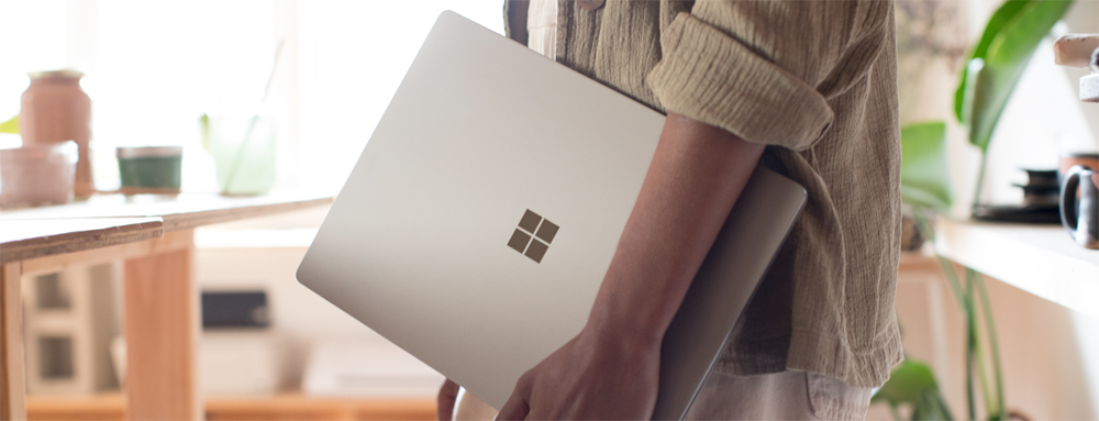 Person at home carrying a closed Microsoft Surface Laptop at their side